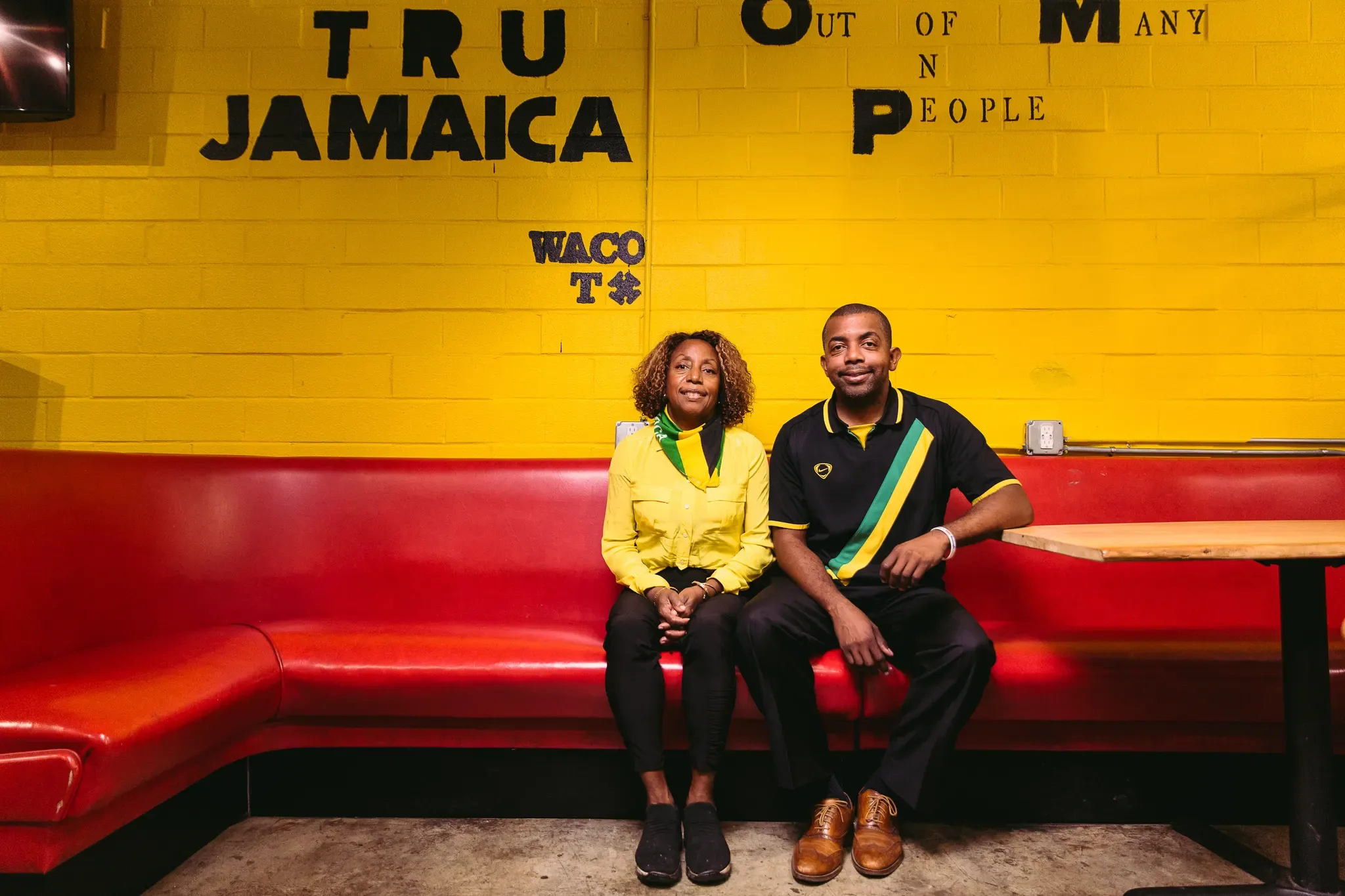 The Tru Jamaica owners seated on the red booth below the yellow Tru Jamaica, Out of Many One People, Waco TX wall