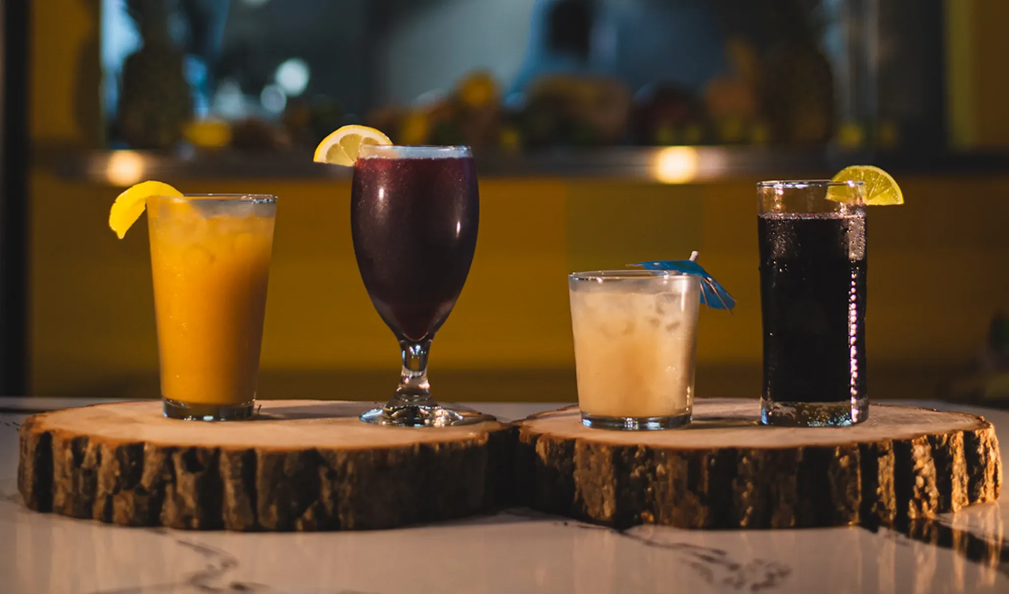 Four fresh Jamaican juices with lemon wedges on wood rounds, glowing in front of the yellow wall